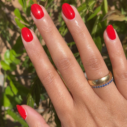 Hand with nails painted in glossy cherry red Lollipop quick dry polish wearing gold and blue gemstone rings outdoors