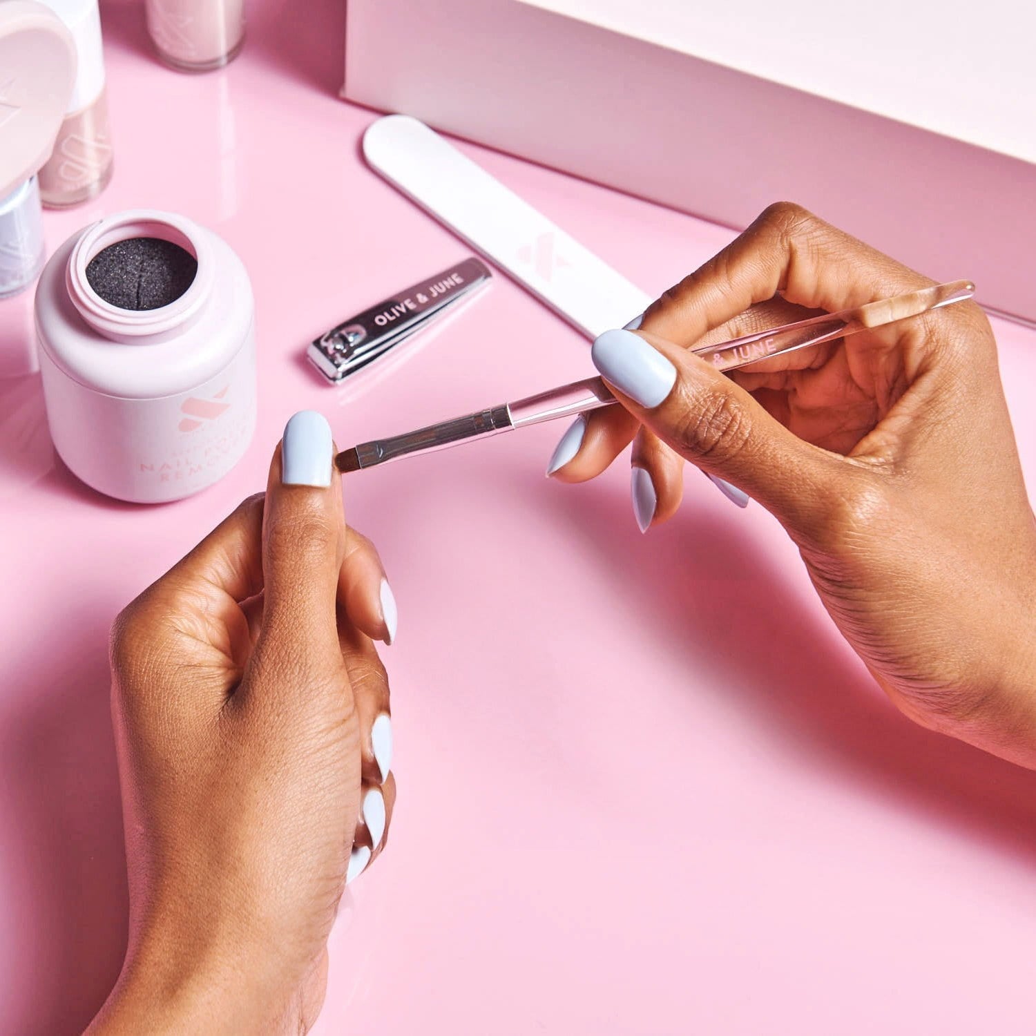 Hands holding the Clean Up Brush cleaning a nail tool with nail care items on a pink surface for manicures